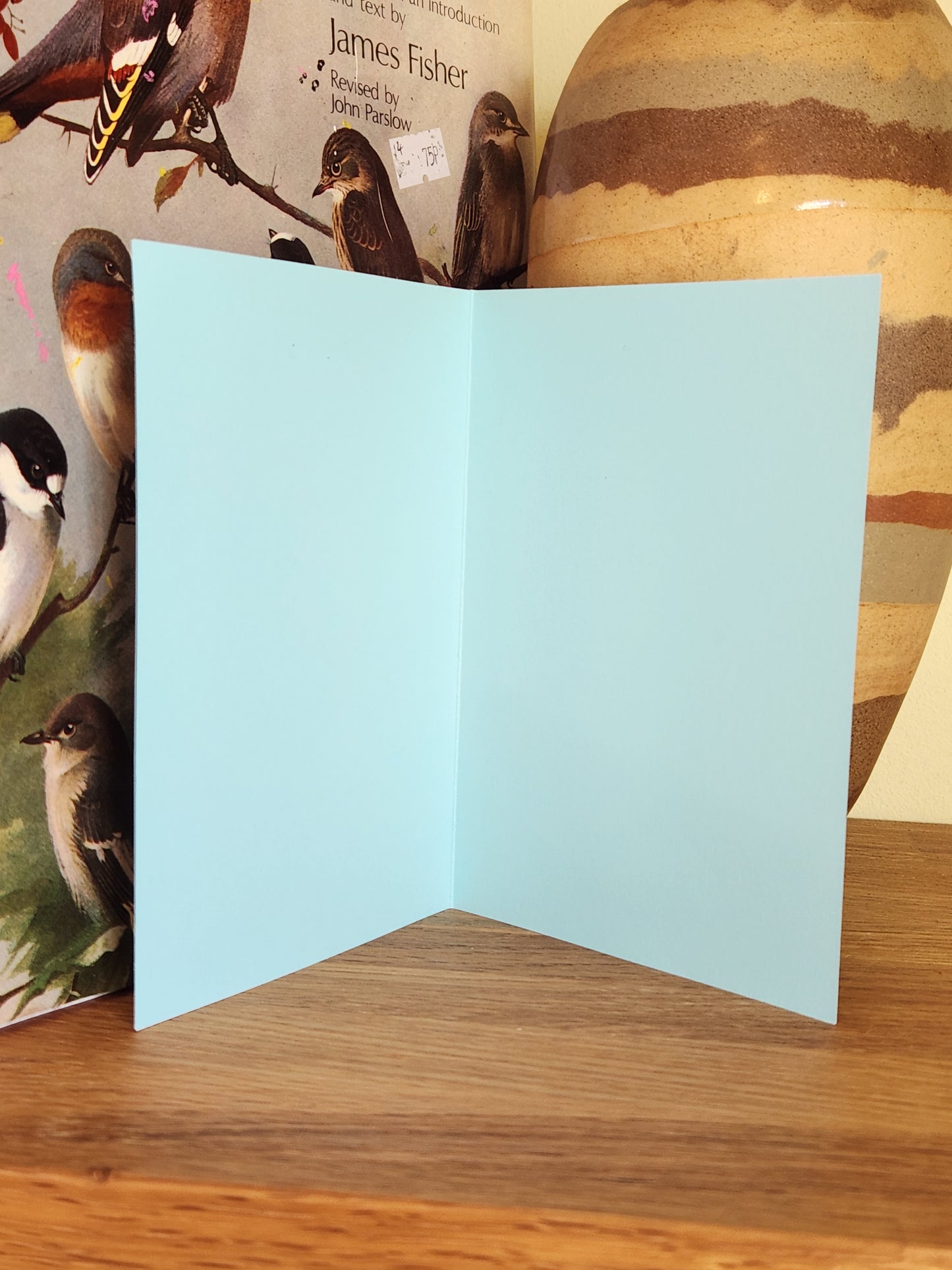 The blank inside of a light blue greeting card standing open on top of a wooden surface with a ceramic vase in the background.