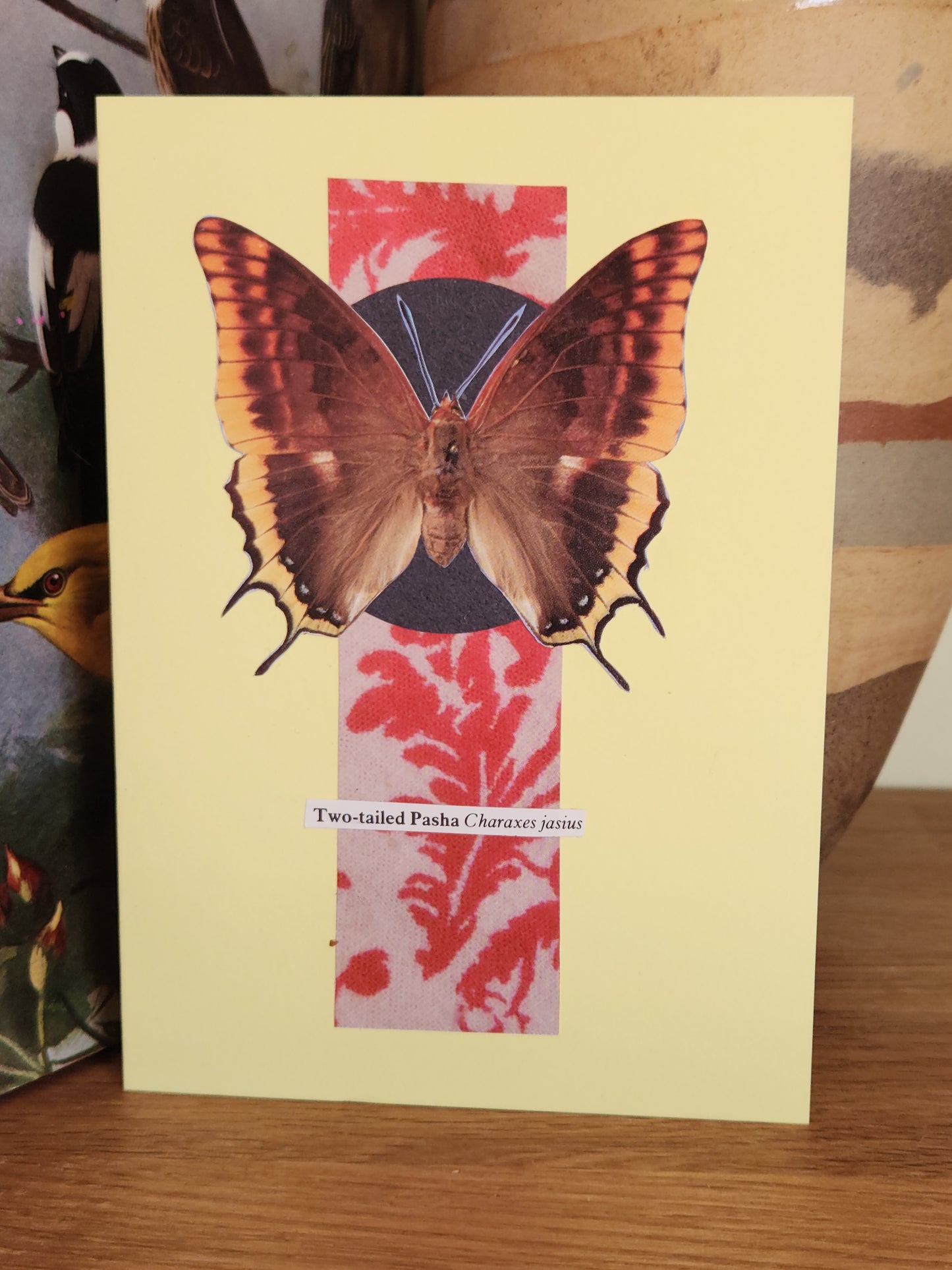 A pale yellow greeting card featuring a collage of a butterfly over red and white abstract collage panels, photographed standing on a wooden surface.