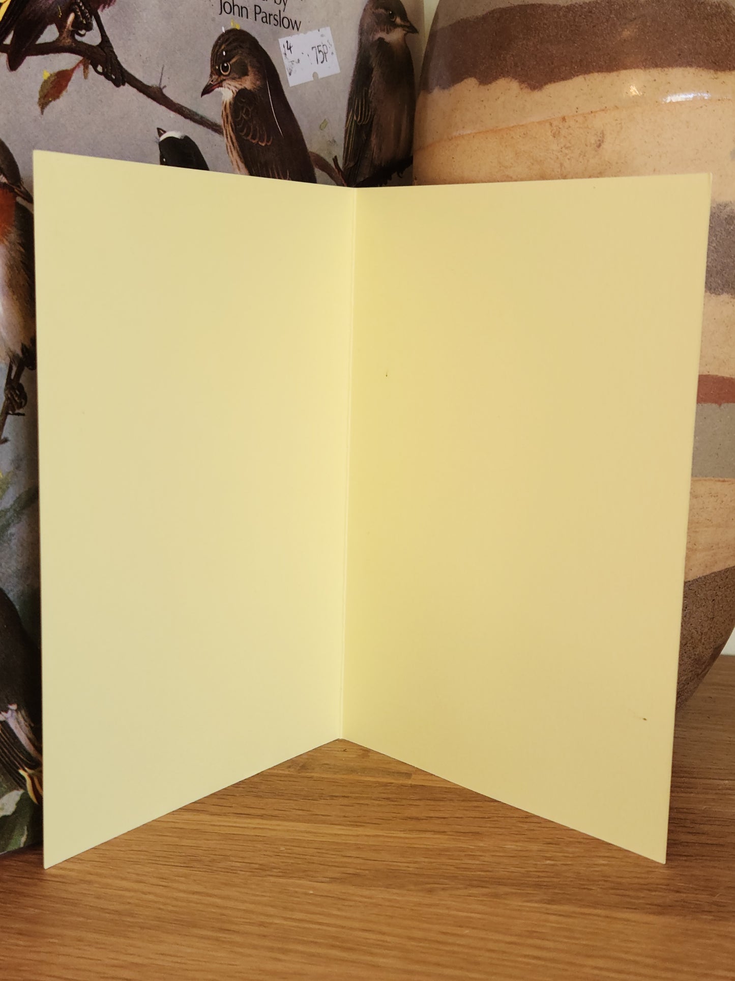 The blank inside of a pale yellow greeting card standing open on a wooden surface, with a striped ceramic vase in the background