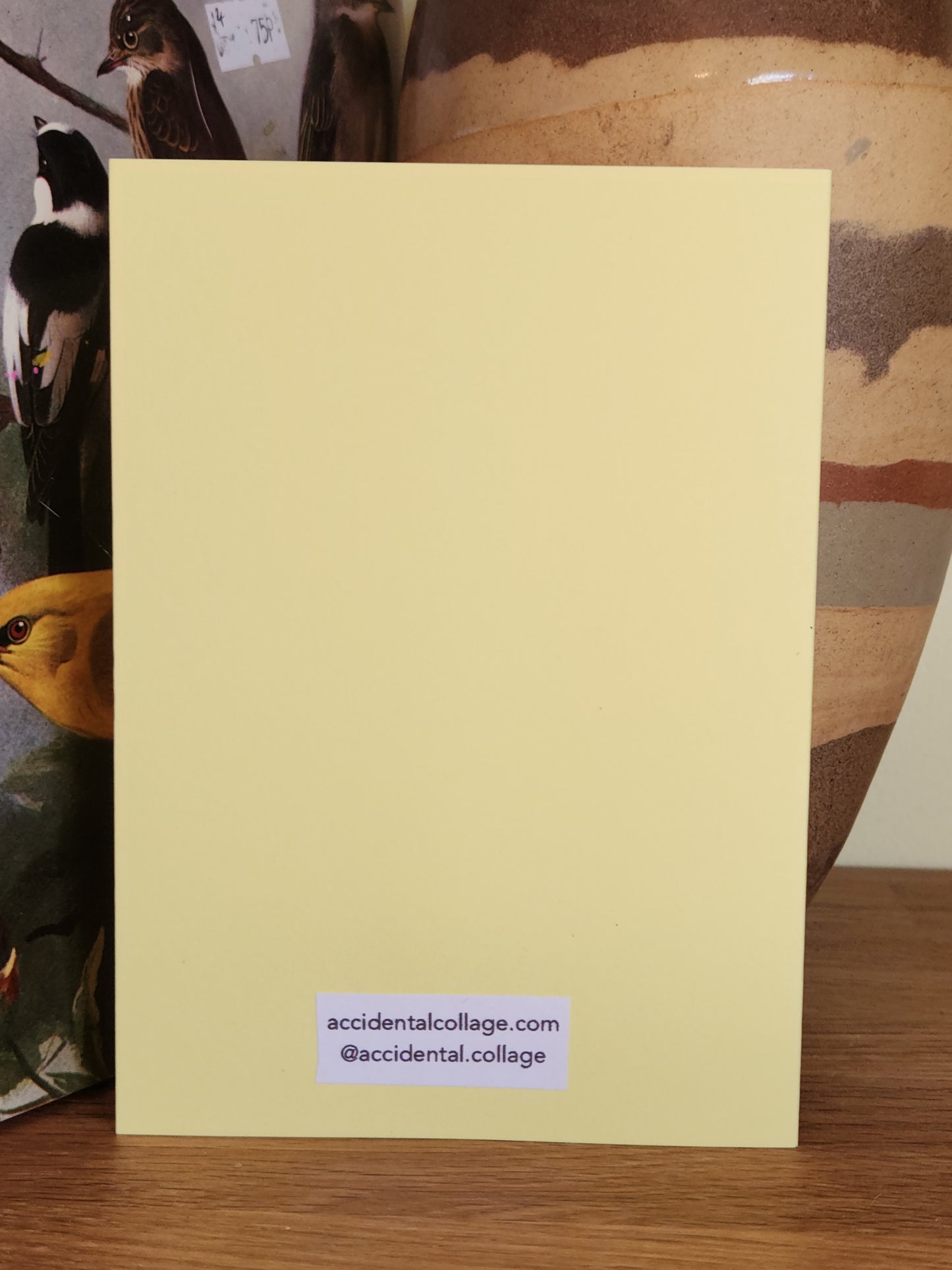 The back of a pale yellow greeting card standing upright on a wooden surface, with a small printed label near the bottom reading accidentalcollage.com and @accidental collage.