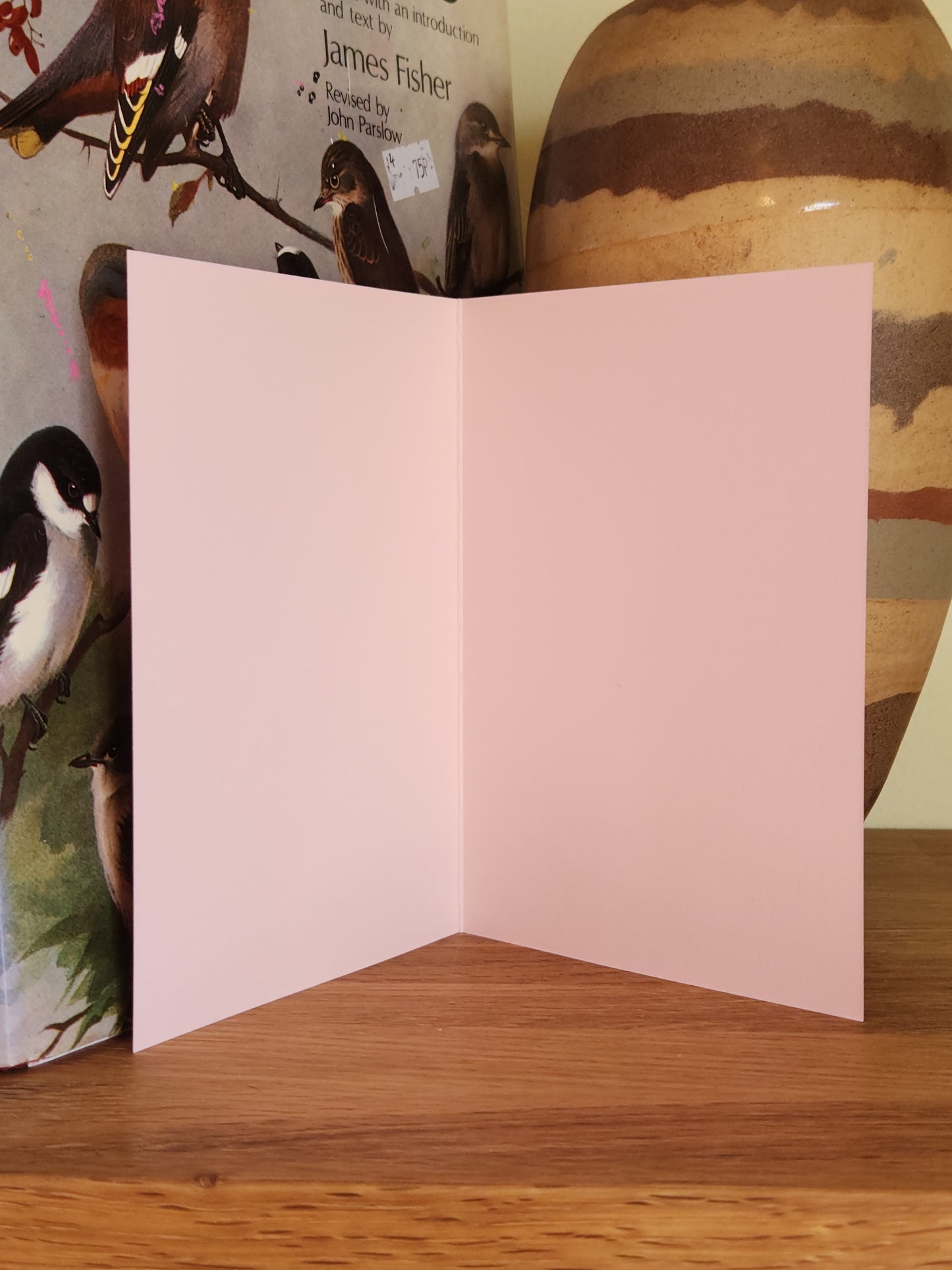 The blank, inside of a pink folded greeting card standing open on a wooden tabletop, with a vase in the background.