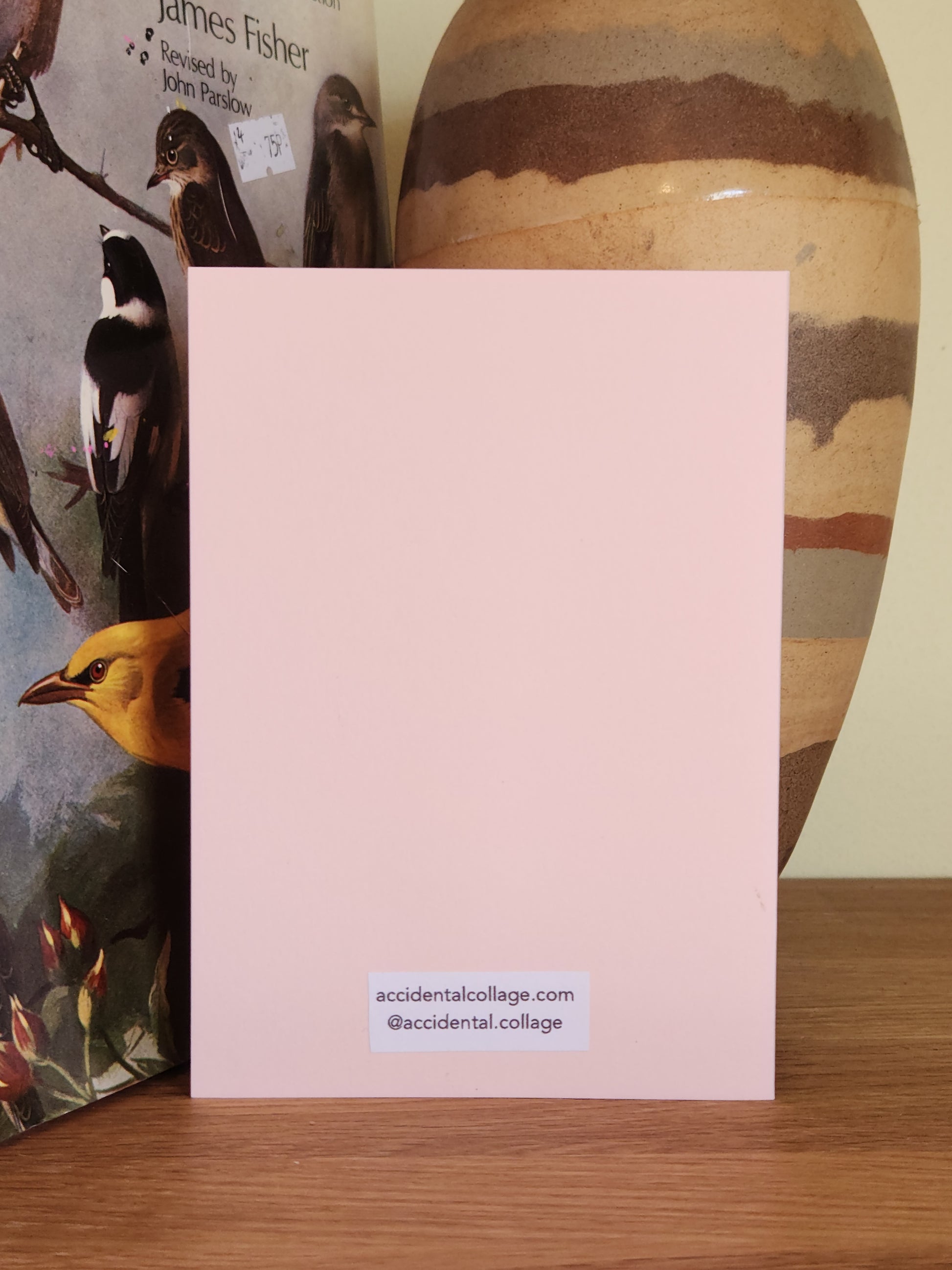 The back of a pale pink greeting card standing upright on a wooden surface, with a small label at the bottom reading 'accidentalcollage.com @accidental.collage'.