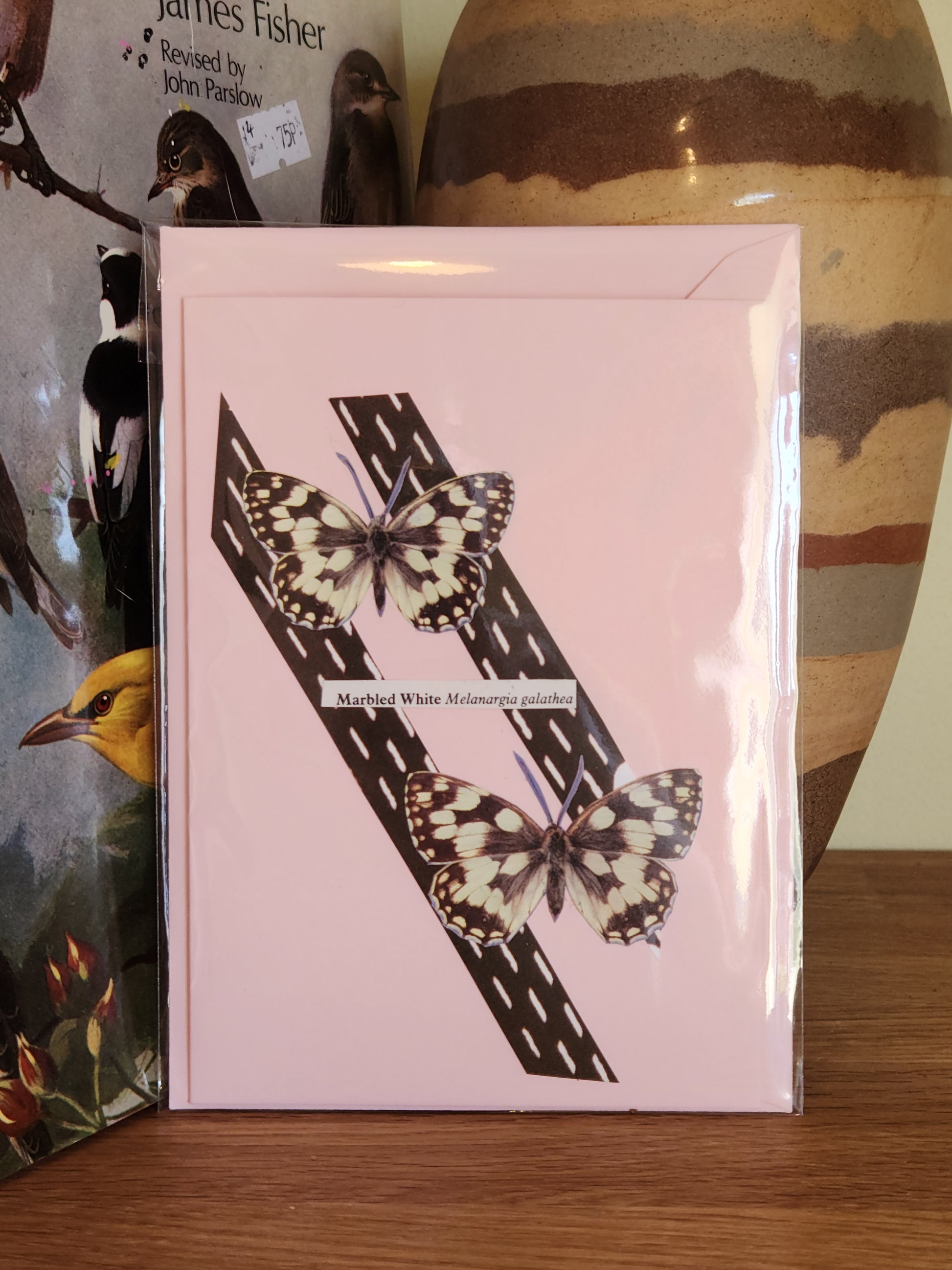 A pale pink greeting card featuring a collage of two black and white butterflies on diagonal black sections, packaged in a plastic sleeve with a matching envelope, displayed on a wooden surface with a striped ceramic vase in the background.