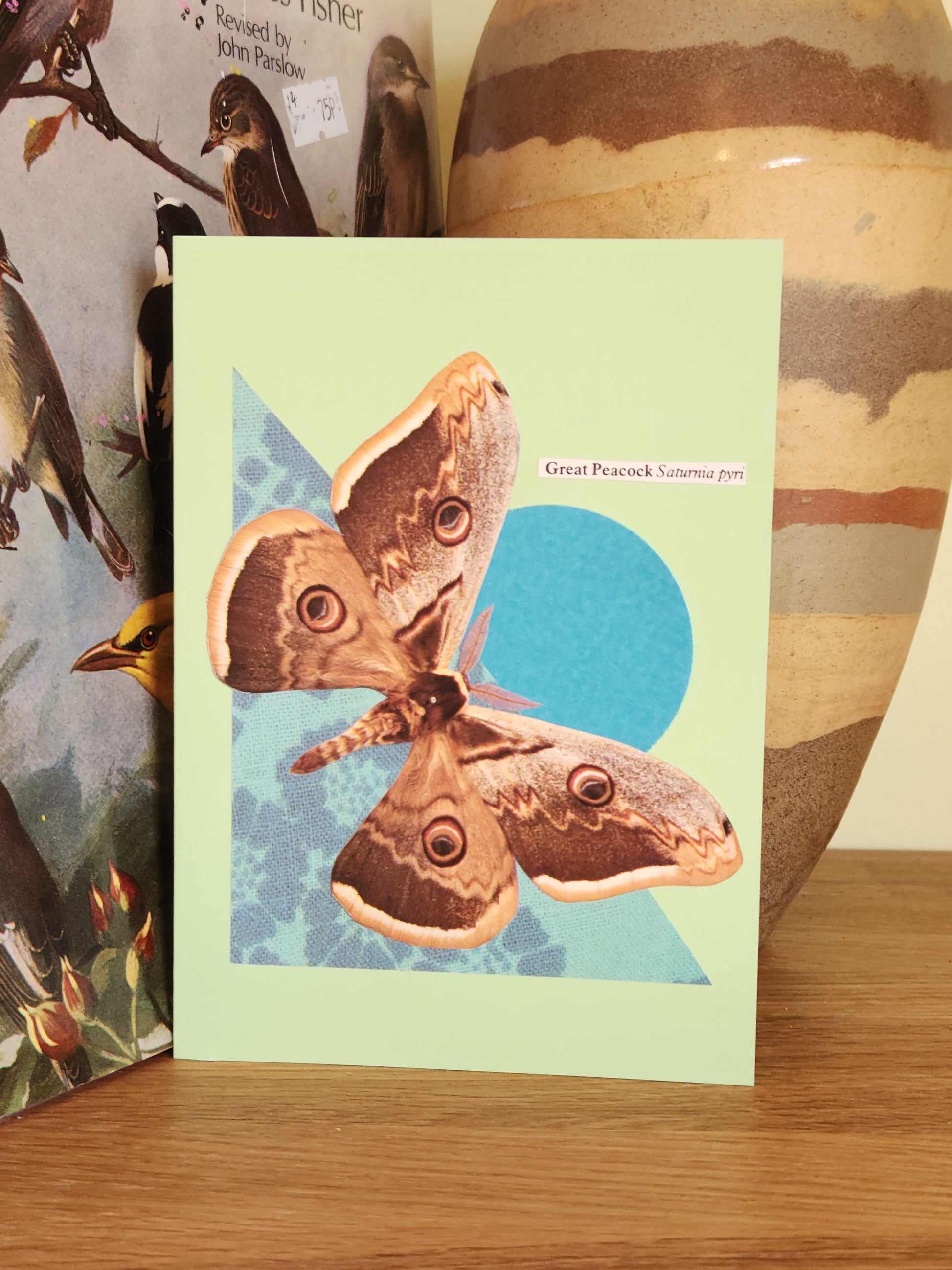 Pale green greeting card with a collage of a Great Peacock moth on a teal circle and teal patterned triangle, on a wooden surface, in front of a striped ceramic vase