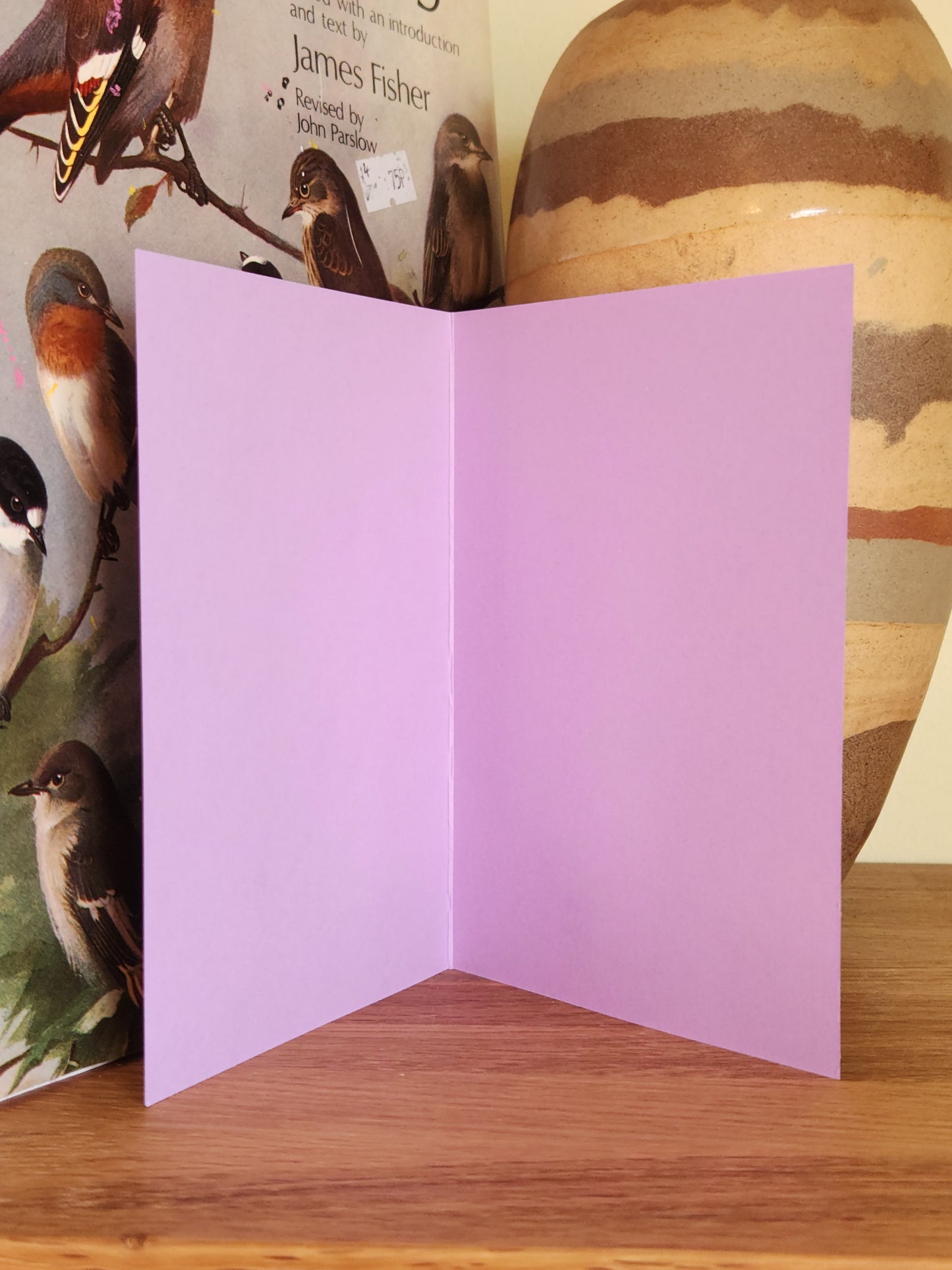 The blank inside of a lavender greeting card standing open on a wooden surface, with a striped ceramic vase in the background.