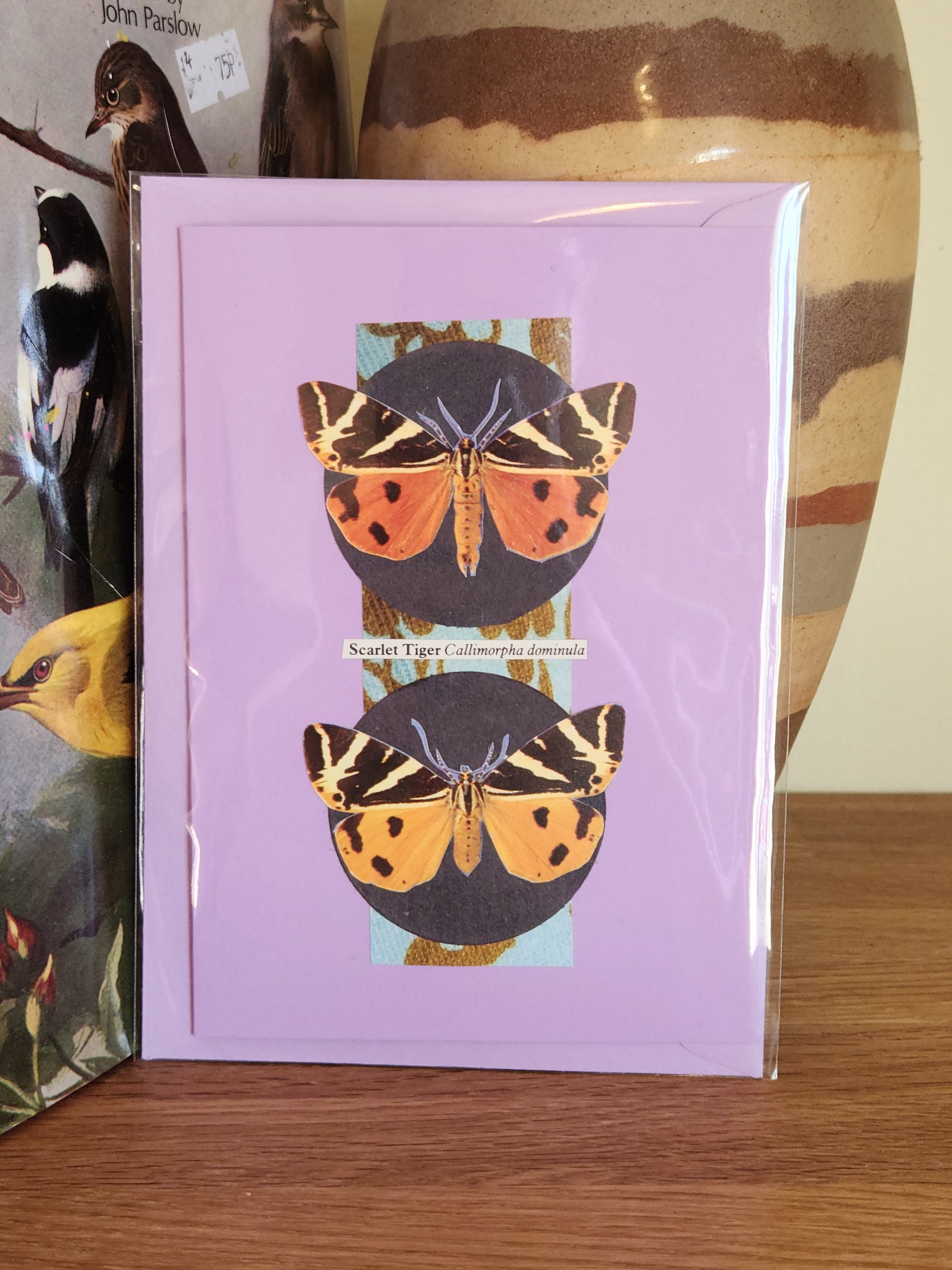 A lavender greeting card featuring a collage of two scarlet tiger moths, packaged in a plastic sleeve with a matching envelope, standing on a wooden surface with a striped ceramic vase in the background