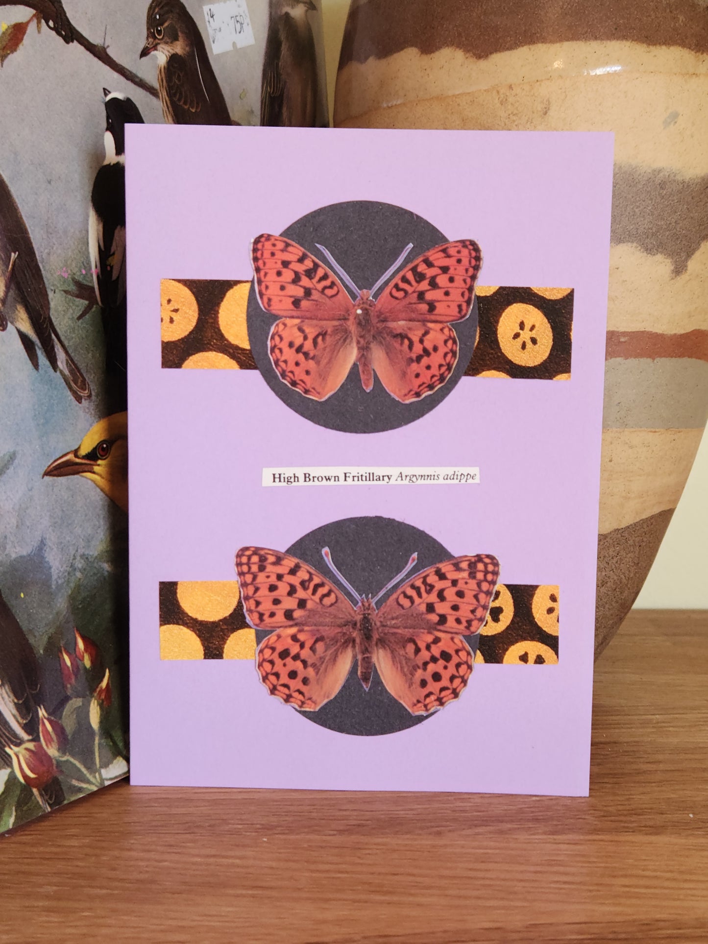A lavender greeting card featuring a collage of two orange and black butterflies on a yellow spotted background, resting on a wooden surface witha ceramic vase in the background.