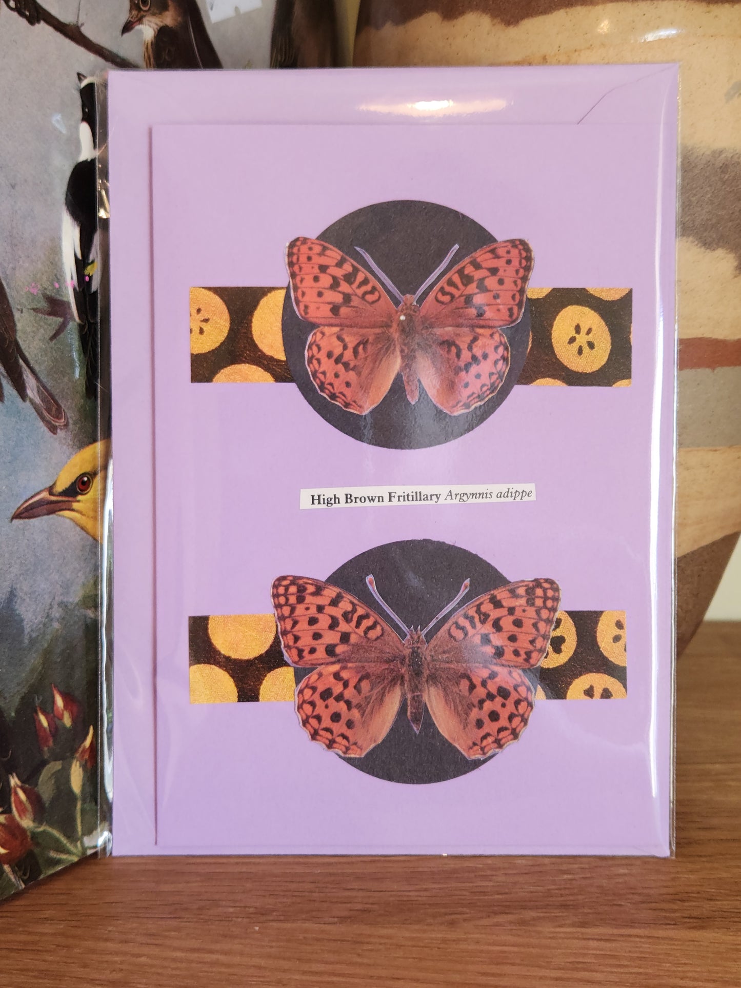 A lavender greeting card featuring a collage of two butterfles with yellow patterned horizontal bands. The card is sealed in plastic packaging with a matching envelope and photographed standing on a wooden surface.