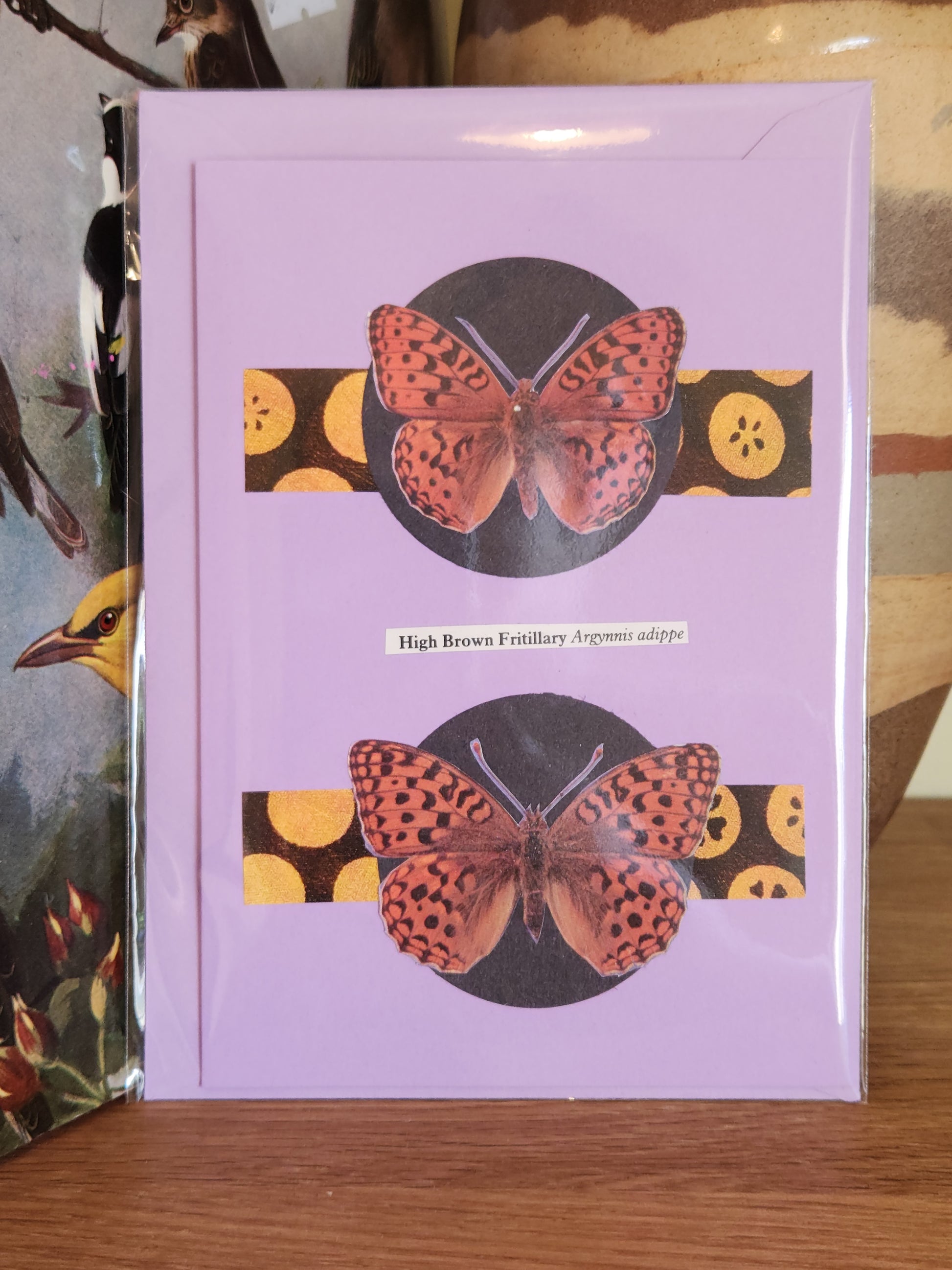 A lavender greeting card featuring a collage of two butterfles with yellow patterned horizontal bands. The card is sealed in plastic packaging with a matching envelope and photographed standing on a wooden surface.