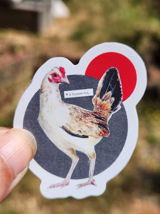 A die-cut sticker featuring an image of a white chicken with red comb and wattle, a large red circle, and a dark gray oval background, held by a person's fingers against a blurred outdoor background.
