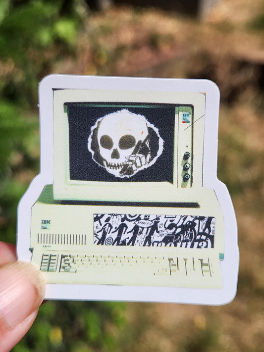 A hand holding a die-cut sticker featuring a vintage computer screen with a skull graphic and patterned side panels, against a blurred outdoor background.