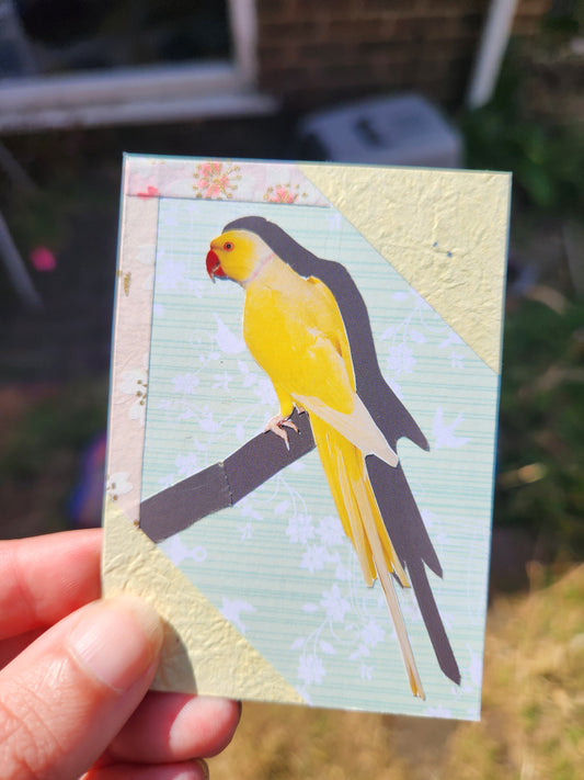A hand holding a small decorative card featuring a yellow bird perched on a branch, with a pale green floral background and textured cream corner sections.