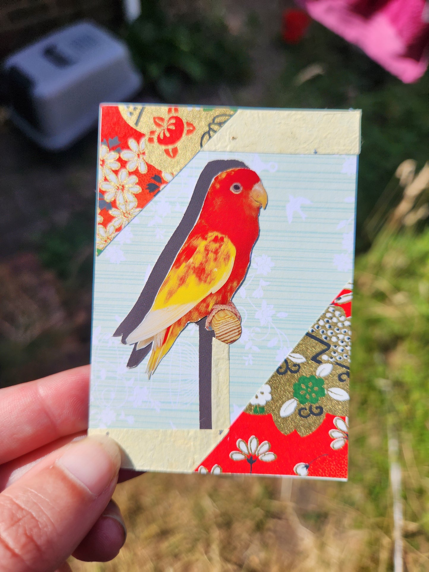 A hand holding a small rectangular decorative card featuring a colorful bird collage with patterned paper sections, photographed outdoors with blurred grass and ground in the background.